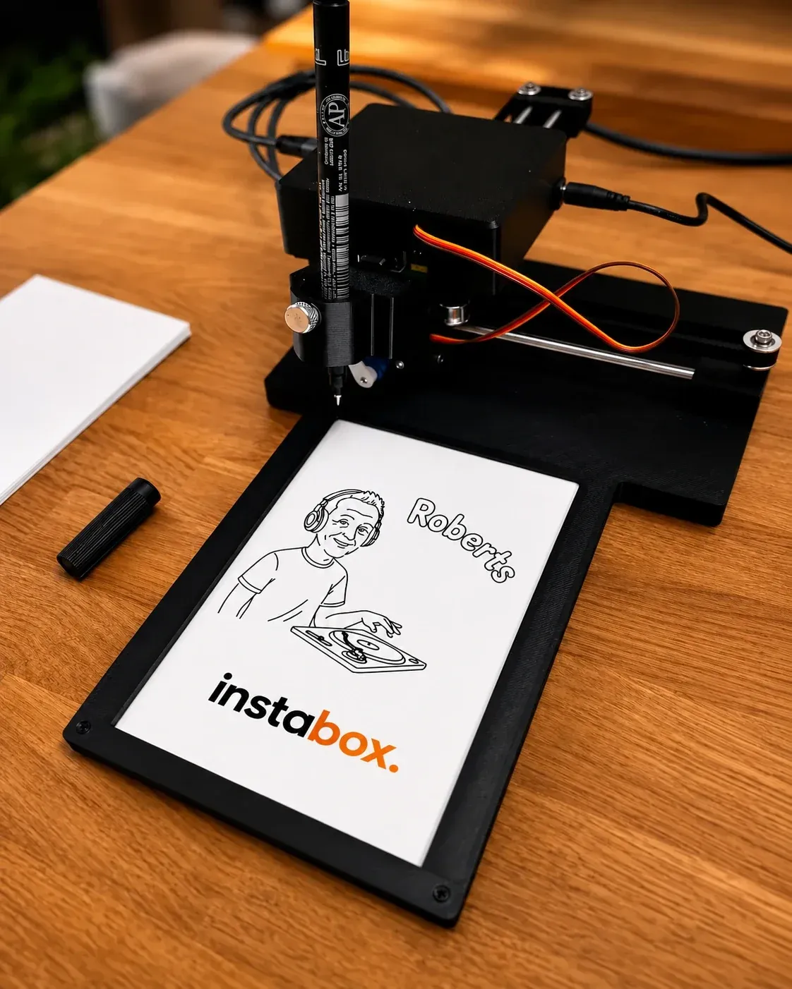Zīmēšanas robots ar pildspalvu uzzīmējis DJ stila karikatūru ar Instabox logo